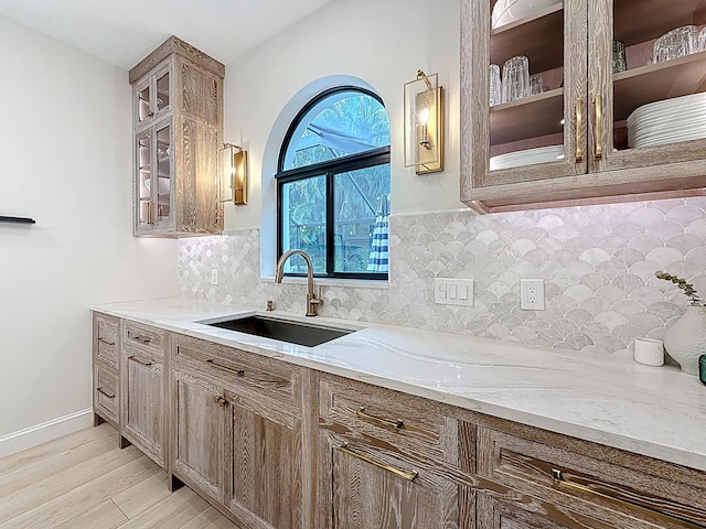 kitchen with decorative backsplash, a sink, light stone counters, light wood finished floors, and glass insert cabinets