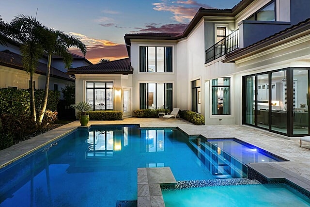 back of house at dusk with a patio, a balcony, stucco siding, an outdoor pool, and a tiled roof