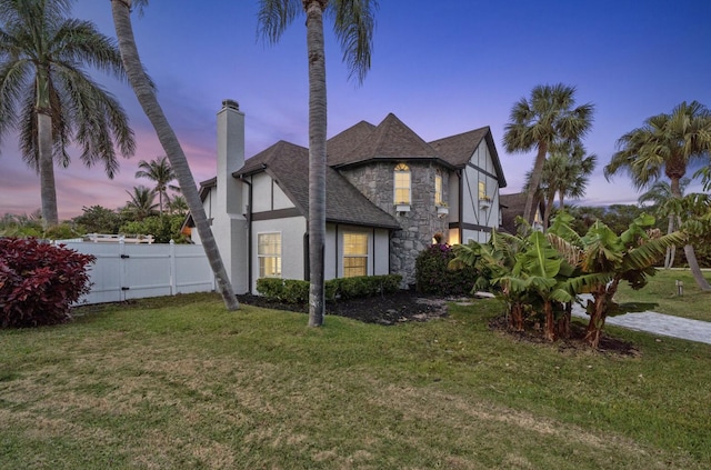 121 Queen Eugenia Ct, Hutchinson Island, FL 34949 - property exterior at dusk with roof with shingles, a chimney, stone siding, a yard, and fence