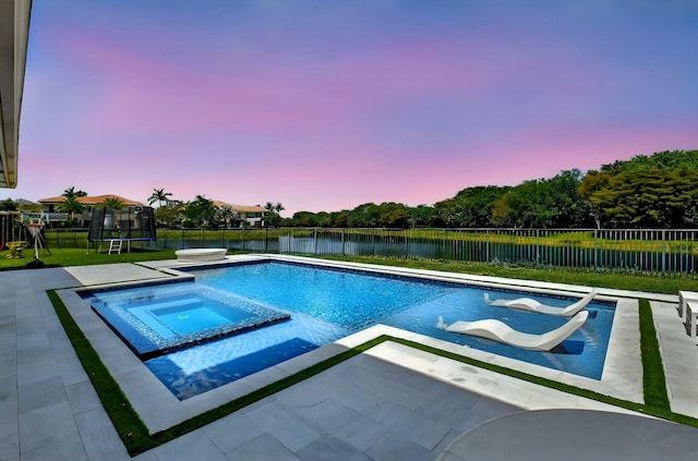 pool at dusk featuring a trampoline, a patio area, a fenced backyard, a water view, and a pool with connected hot tub