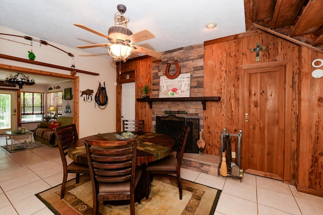 dining area with wooden walls, light tile patterned flooring, ceiling fan, and a stone fireplace