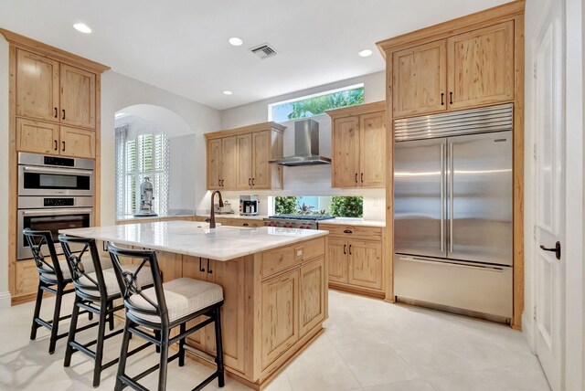kitchen featuring arched walkways, a breakfast bar, appliances with stainless steel finishes, wall chimney range hood, and a sink