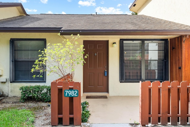 entrance to property featuring roof with shingles, stucco siding, and fence