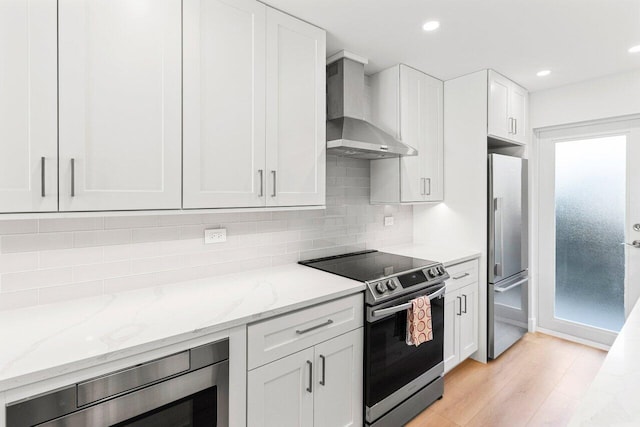 kitchen with tasteful backsplash, white cabinets, light stone countertops, wall chimney exhaust hood, and appliances with stainless steel finishes