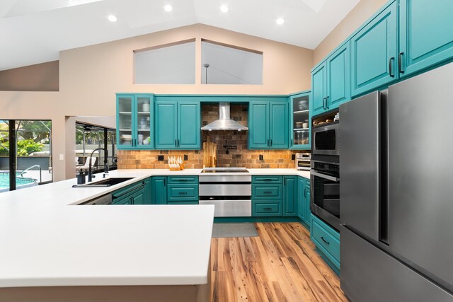 kitchen with light wood-style floors, wall chimney range hood, a sink, appliances with stainless steel finishes, and vaulted ceiling