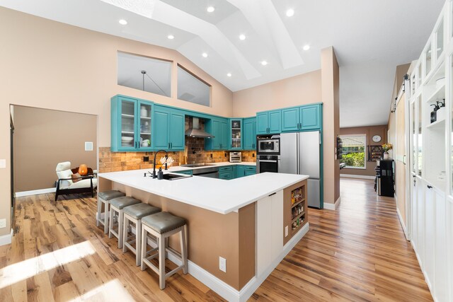 kitchen featuring appliances with stainless steel finishes, a sink, wall chimney exhaust hood, a peninsula, and blue cabinets