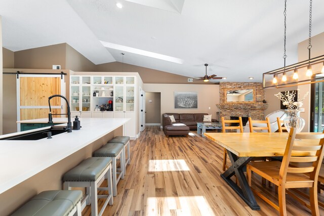 dining room with ceiling fan, a barn door, light wood finished floors, a fireplace, and lofted ceiling with skylight
