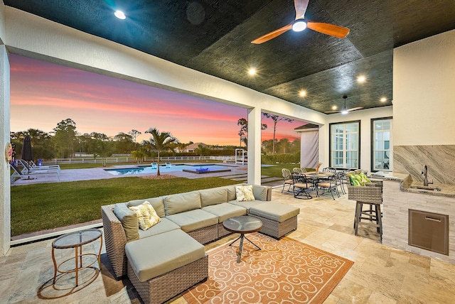patio terrace at dusk featuring an outdoor kitchen, outdoor lounge area, a ceiling fan, a sink, and an outdoor pool