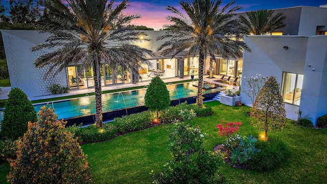 back of property at dusk with an outdoor pool, a patio area, a lawn, and stucco siding