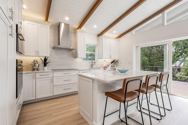 kitchen featuring lofted ceiling with beams, white cabinets, tasteful backsplash, a kitchen bar, and wall chimney range hood
