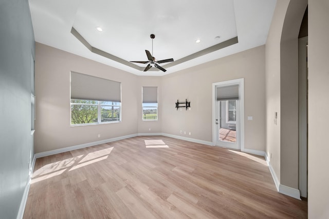 empty room with a raised ceiling, ceiling fan, light wood-style floors, baseboards, and arched walkways