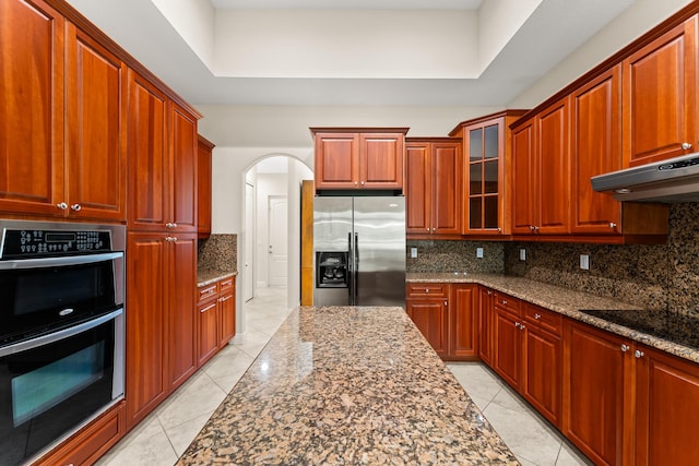 kitchen featuring dark stone counters, decorative backsplash, arched walkways, stainless steel appliances, and under cabinet range hood