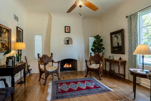 living area with visible vents, a ceiling fan, a textured wall, hardwood / wood-style floors, and a fireplace with flush hearth