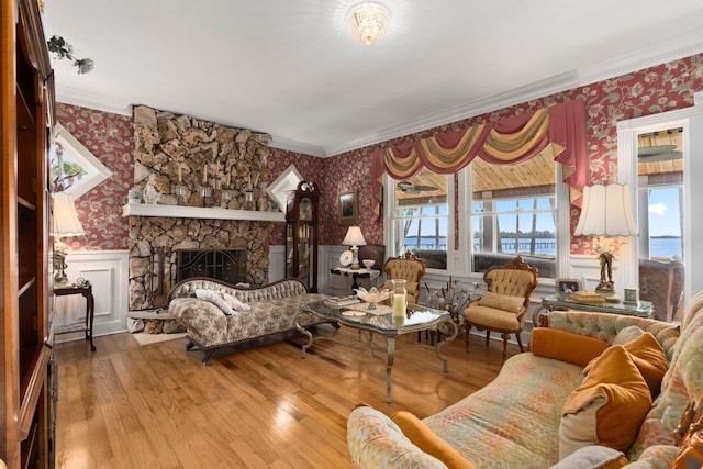 living room with wainscoting, wallpapered walls, crown molding, hardwood / wood-style flooring, and a stone fireplace