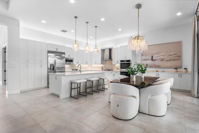 kitchen with built in appliances, wall chimney range hood, a center island with sink, a chandelier, and recessed lighting
