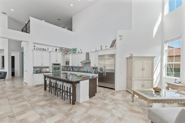 kitchen with a breakfast bar, stainless steel appliances, wall chimney exhaust hood, a kitchen island with sink, and a high ceiling