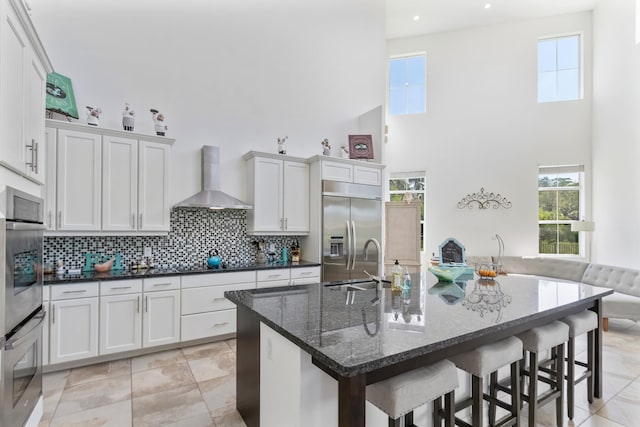 kitchen featuring wall chimney range hood, a breakfast bar area, a sink, appliances with stainless steel finishes, and a high ceiling