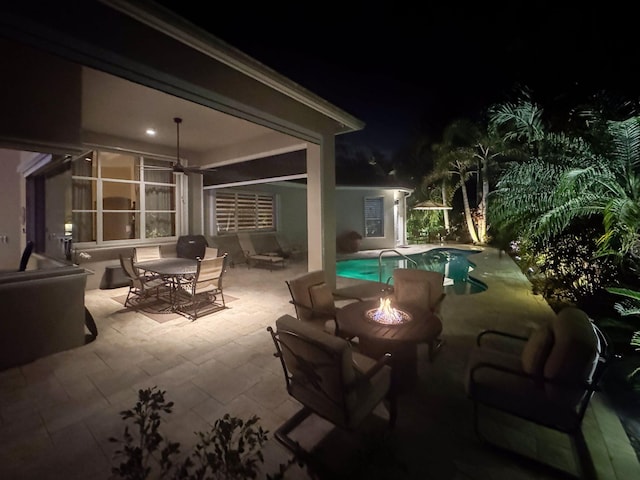 patio at night featuring outdoor dining area, a ceiling fan, a fire pit, and an outdoor pool
