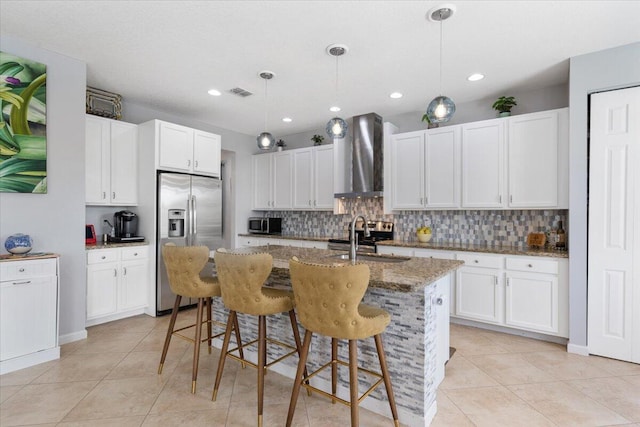 kitchen featuring stainless steel refrigerator with ice dispenser, wall chimney range hood, a sink, decorative backsplash, and light stone counters