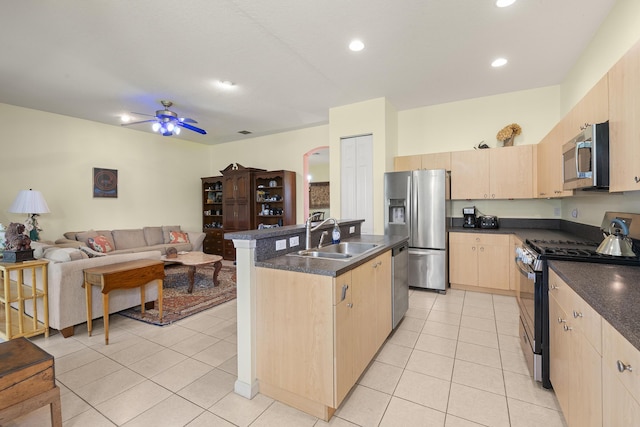kitchen featuring light brown cabinetry, appliances with stainless steel finishes, dark countertops, a sink, and open floor plan