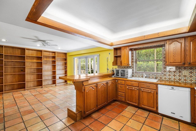 kitchen featuring a sink, white dishwasher, a raised ceiling, backsplash, and a peninsula