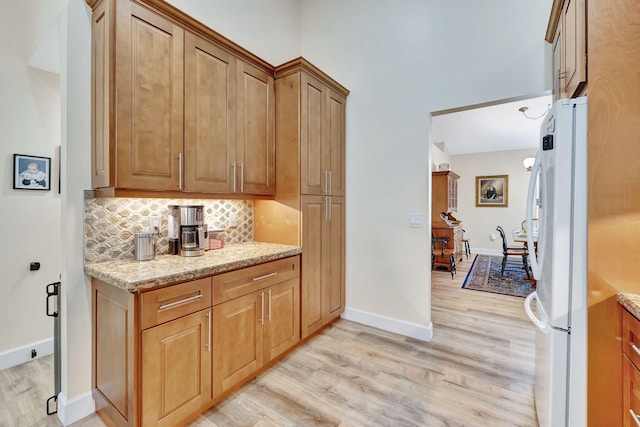kitchen featuring light stone counters, decorative backsplash, light wood-style flooring, freestanding refrigerator, and baseboards
