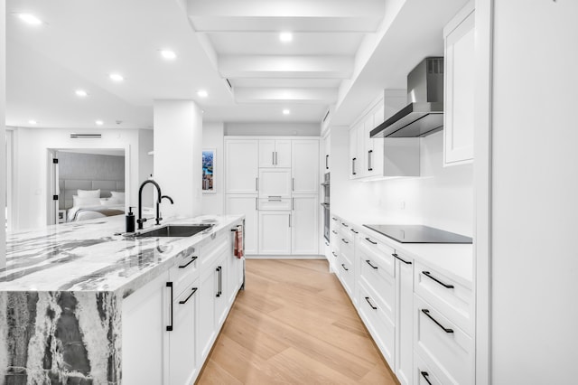 kitchen with black electric stovetop, light wood-style flooring, wall chimney range hood, a sink, and white cabinetry