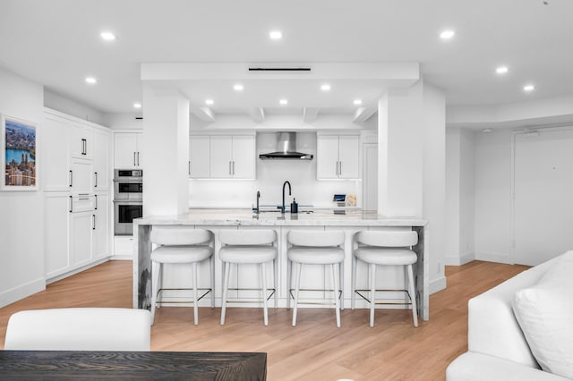 kitchen featuring light wood-type flooring, stainless steel double oven, wall chimney exhaust hood, and a kitchen breakfast bar