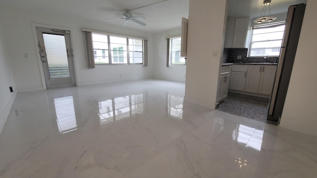 unfurnished living room featuring healthy amount of natural light, a ceiling fan, baseboards, and light marble finish flooring