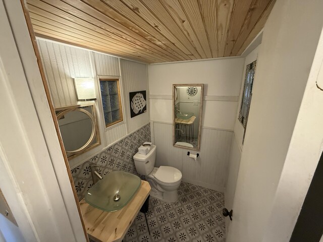 bathroom with a sink, wooden ceiling, tile patterned floors, a wainscoted wall, and toilet