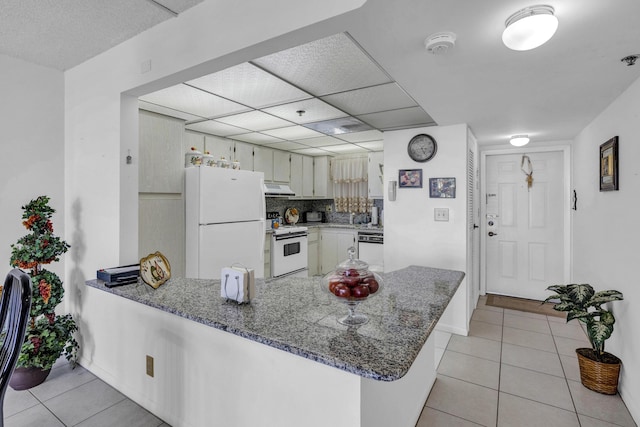 kitchen with a peninsula, white appliances, under cabinet range hood, stone counters, and light tile patterned floors