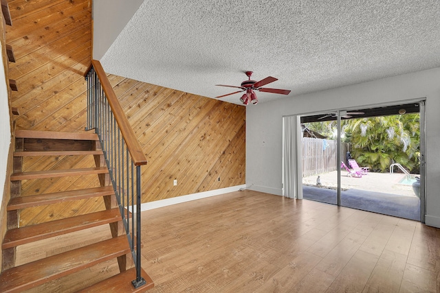 unfurnished living room with wooden walls, a textured ceiling, stairs, a ceiling fan, and baseboards