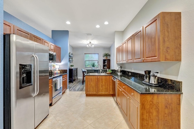 kitchen with appliances with stainless steel finishes, a peninsula, a sink, light tile patterned floors, and dark stone counters