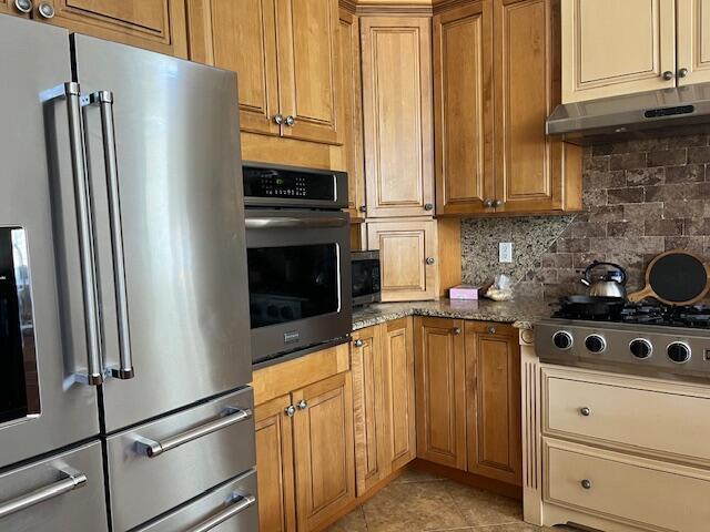 kitchen with decorative backsplash, light tile patterned flooring, dark stone counters, under cabinet range hood, and stainless steel appliances
