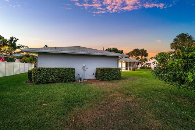 back of house at dusk featuring a lawn, fence, roof with shingles, and stucco siding
