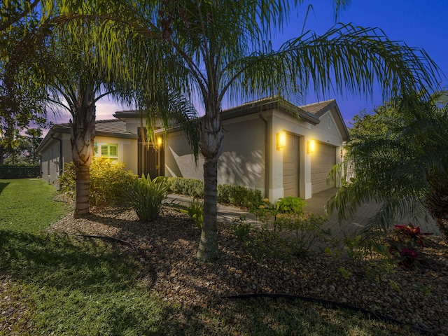 property exterior at dusk with driveway, stucco siding, a tiled roof, an attached garage, and a yard