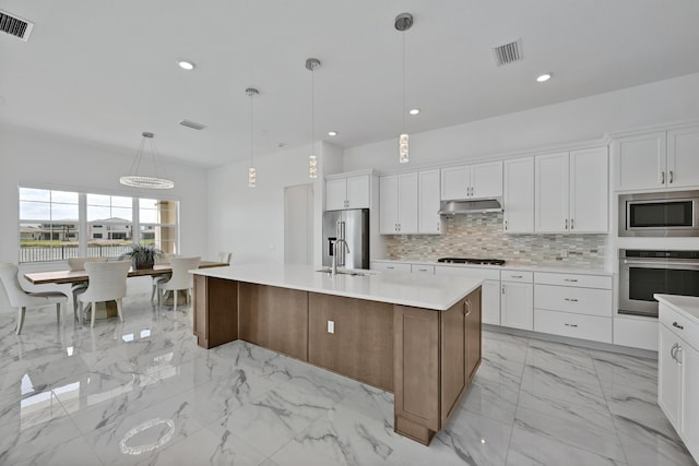 kitchen with appliances with stainless steel finishes, backsplash, under cabinet range hood, visible vents, and a center island with sink
