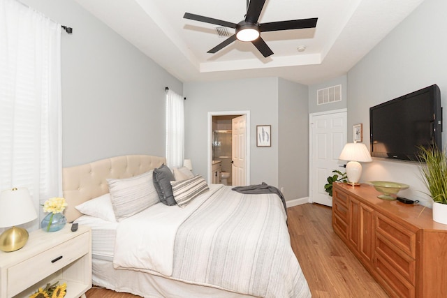 bedroom featuring visible vents, a raised ceiling, baseboards, light wood finished floors, and ensuite bathroom