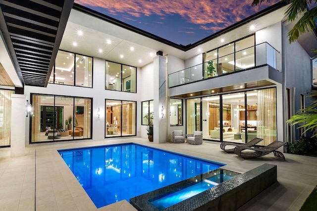 back of house at dusk featuring a balcony, a patio area, an outdoor living space, a pool with connected hot tub, and stucco siding