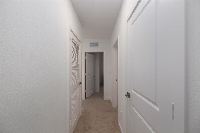 hallway with visible vents, a textured wall, and light colored carpet