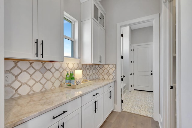 kitchen featuring white cabinets, light stone counters, light wood finished floors, decorative backsplash, and glass insert cabinets