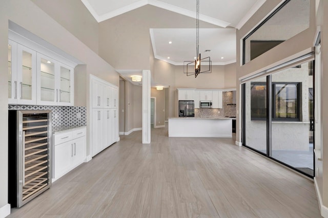 unfurnished living room with crown molding, wine cooler, light wood-style floors, high vaulted ceiling, and a chandelier