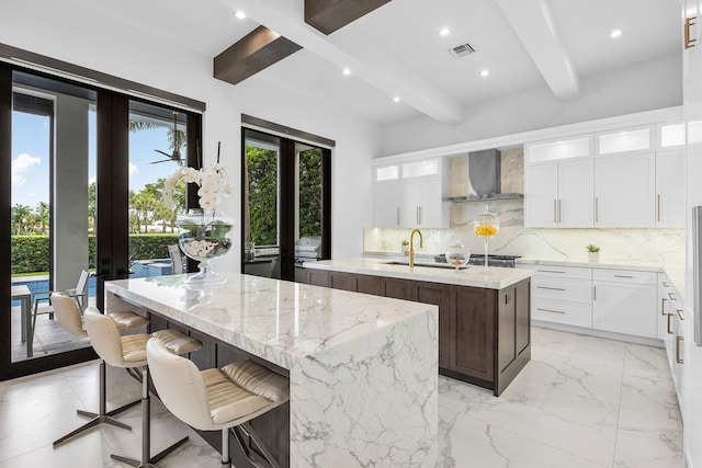 kitchen with an island with sink, wall chimney exhaust hood, a sink, beamed ceiling, and backsplash