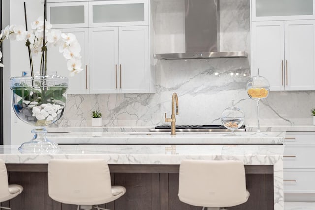 kitchen with wall chimney exhaust hood, white cabinets, a breakfast bar, tasteful backsplash, and light stone countertops