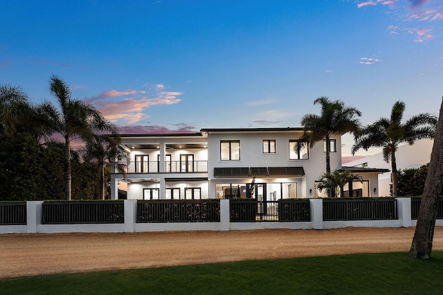 back of house at dusk with a balcony, stucco siding, and a fenced front yard