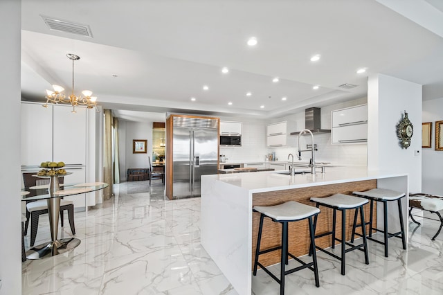 kitchen featuring built in refrigerator, wall chimney exhaust hood, a peninsula, modern cabinets, and a kitchen bar