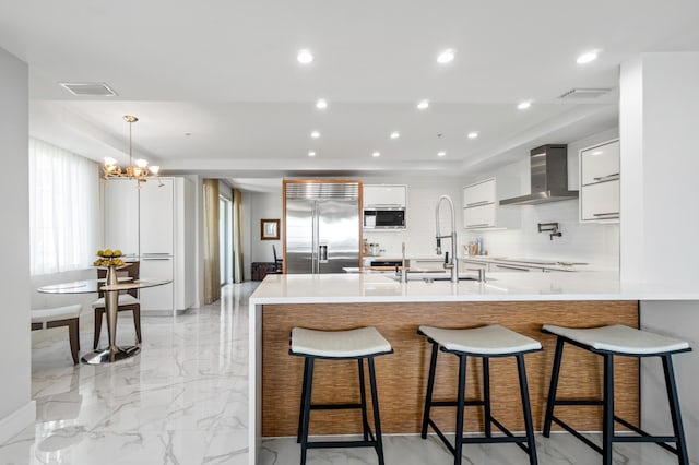 kitchen with stainless steel built in fridge, wall chimney exhaust hood, modern cabinets, a chandelier, and light marble finish floors