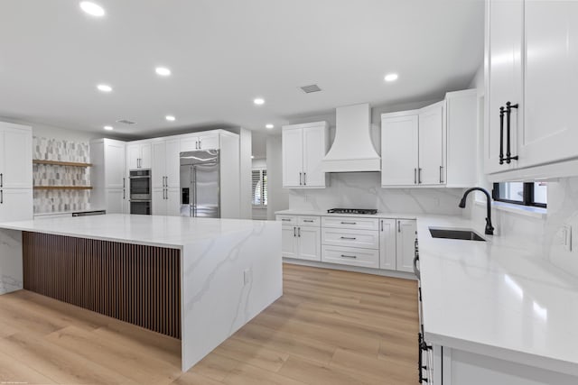 kitchen featuring custom exhaust hood, a sink, appliances with stainless steel finishes, open shelves, and light stone counters