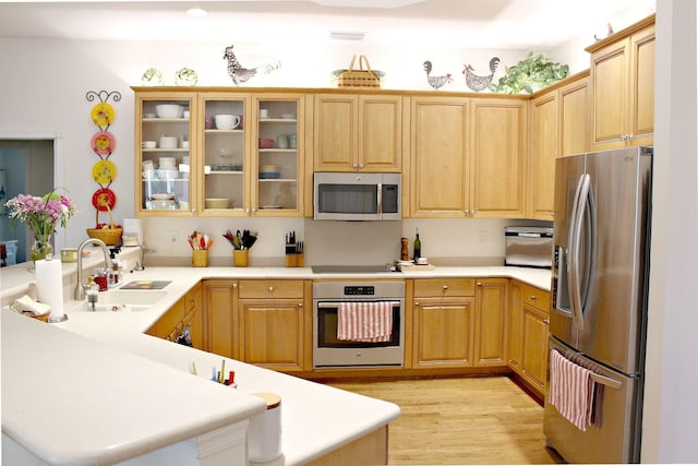 kitchen with appliances with stainless steel finishes, a sink, a peninsula, light wood-style floors, and glass insert cabinets