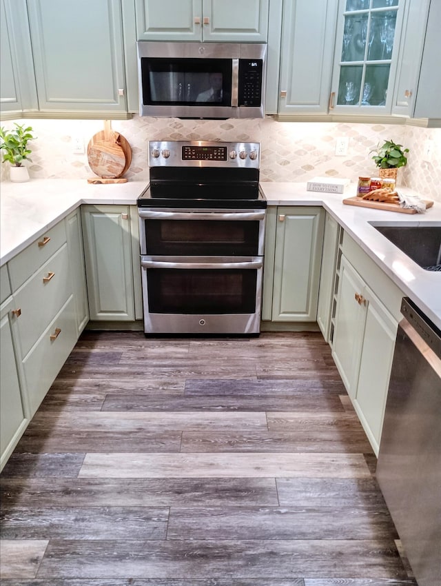 kitchen with glass insert cabinets, wood finished floors, light countertops, tasteful backsplash, and appliances with stainless steel finishes